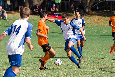 Two teenage boys are both mid stride with the ball between them, one is a Hurlstone player and the other player is wearing orange and black