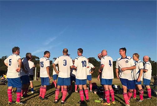 A team of adult men in white, blue and pink-socked uniforms stand with their back to the camera listening to someone talk, perhaps at half time