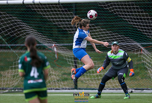 A woman is jumping up to head a ball right in front of goal, the goalie is poised ready to defend and is staring intently at the player