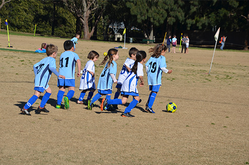 Sixvery young boys and girls run after one girl at the front of the pack who has the ball during a game