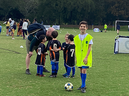 A young male mini-ref wearing a hi-vis vest over his blue and white kit, smiles at camera, small kids are huddled on pitch in background listening to a Dad or coacha